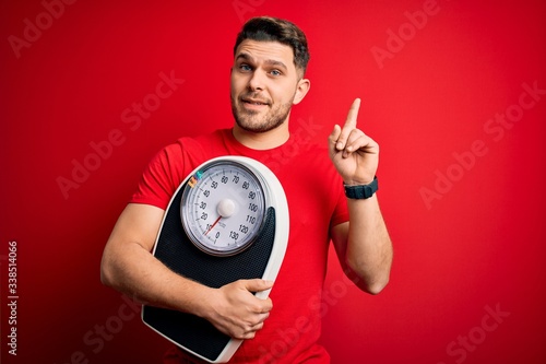 Young fitness man with blue eyes holding scale dieting for healthy weight over red background surprised with an idea or question pointing finger with happy face, number one