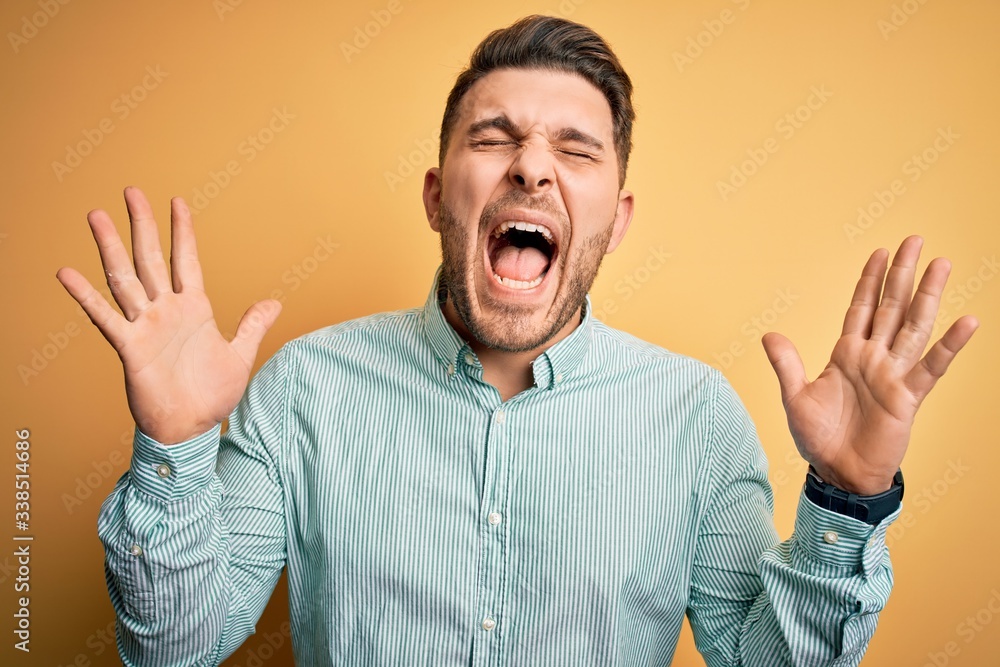 Young business man with blue eyes wearing elegant green shirt over yellow background celebrating mad and crazy for success with arms raised and closed eyes screaming excited. Winner concept Young business man with blue eyes wearing elegant green shirt over yellow background celebrating mad and crazy for success with arms raised and closed eyes screaming excited. Winner concept