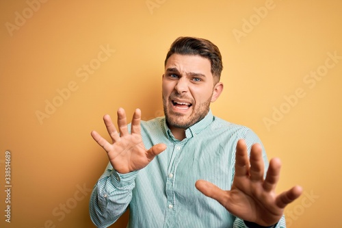 Young business man with blue eyes wearing elegant green shirt over yellow background afraid and terrified with fear expression stop gesture with hands, shouting in shock. Panic concept.