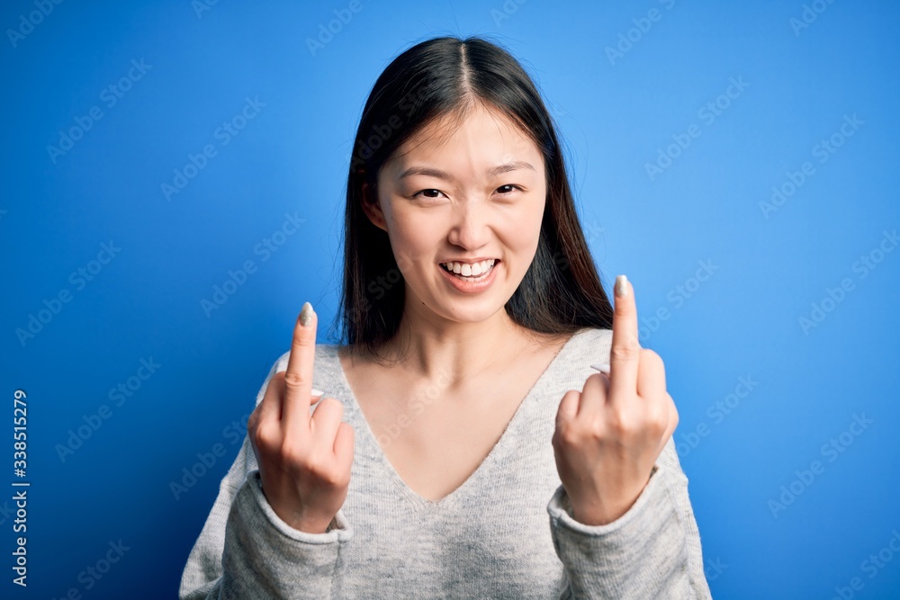 Young beautiful asian woman wearing casual sweater standing over blue isolated background Showing middle finger doing fuck you bad expression, provocation and rude attitude. Screaming excited Young beautiful asian woman wearing casual sweater standing over blue isolated background Showing middle finger doing fuck you bad expression, provocation and rude attitude. Screaming excited