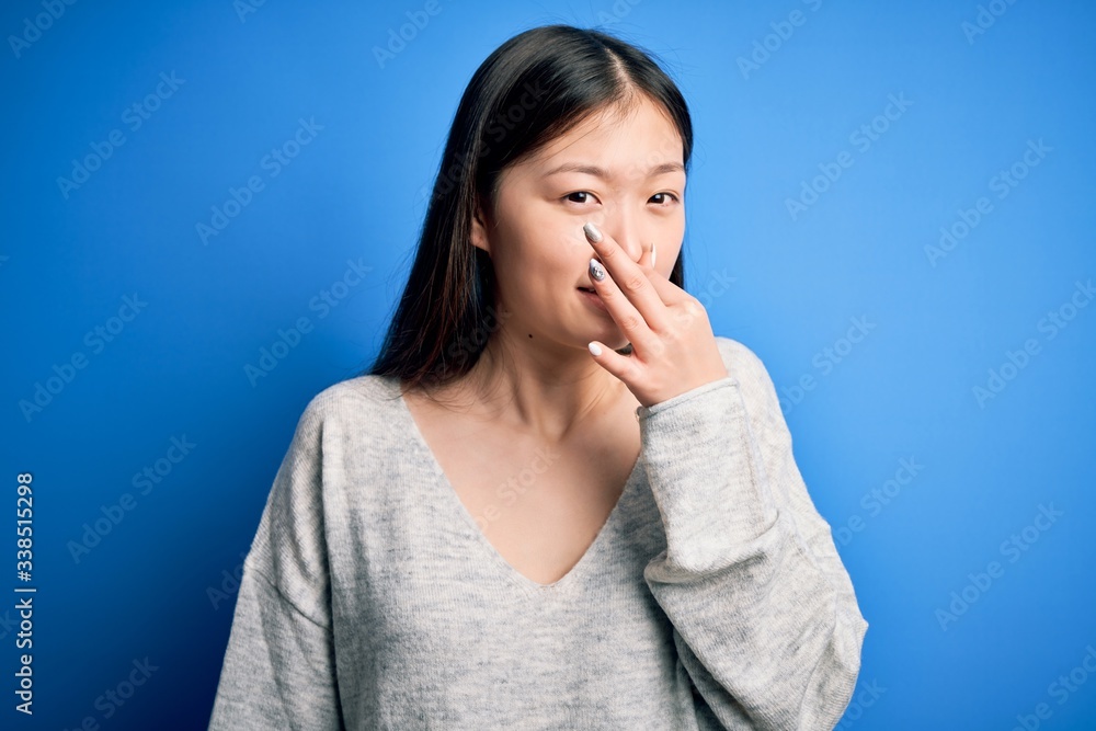 Young beautiful asian woman wearing casual sweater standing over blue isolated background smelling something stinky and disgusting, intolerable smell, holding breath with fingers on nose. Bad smell Young beautiful asian woman wearing casual sweater standing over blue isolated background smelling something stinky and disgusting, intolerable smell, holding breath with fingers on nose. Bad smell
