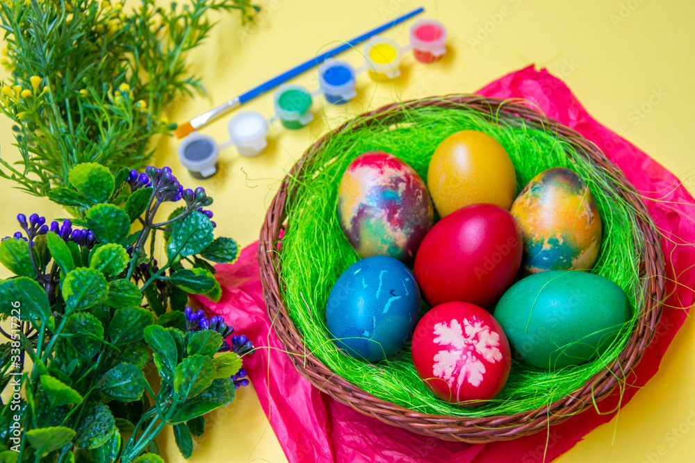 Green grass nest in a basket with colored chicken Easter eggs, multicolored painted eggs,paints and plastic flowers, Easter tradition, celebration concept Green grass nest in a basket with colored chicken Easter eggs, multicolored painted eggs,paints and plastic flowers, Easter tradition, celebration concept
