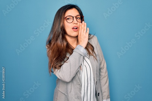 Young hispanic business woman wearing glasses standing over blue isolated background hand on mouth telling secret rumor  whispering malicious talk conversation