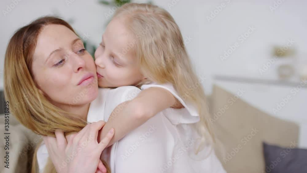 Handheld shot of happy little girl with blond hair hugging her cheerful mother and giving her kiss on cheek Handheld shot of happy little girl with blond hair hugging her cheerful mother and giving her kiss on cheek