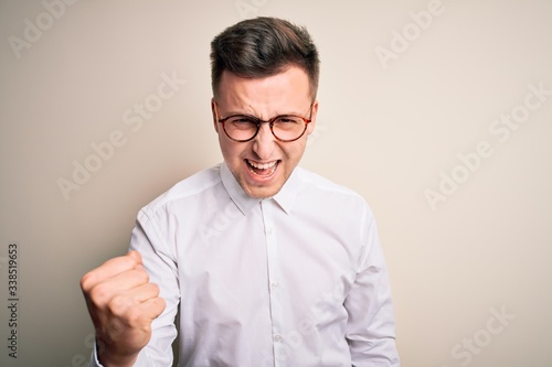 Young handsome business mas wearing glasses and elegant shirt over isolated background angry and mad raising fist frustrated and furious while shouting with anger. Rage and aggressive concept. © Krakenimages.com