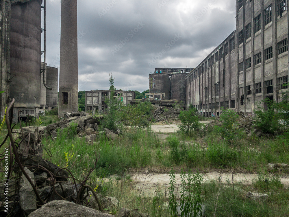 An abandoned, overgrown industrial estate, with tall pipes, ruined buildings, weeds and piles of rubbish