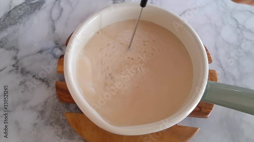 Frothing plant milk for morning latte coffee in an enamel saucepan with electric frother. Top view