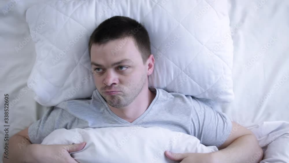 Young man trying to sleep covering ears with pillow. Man lying on bed with covered her ears because of noise. He is angry and looks up at ceiling. Noisy neighbors concept