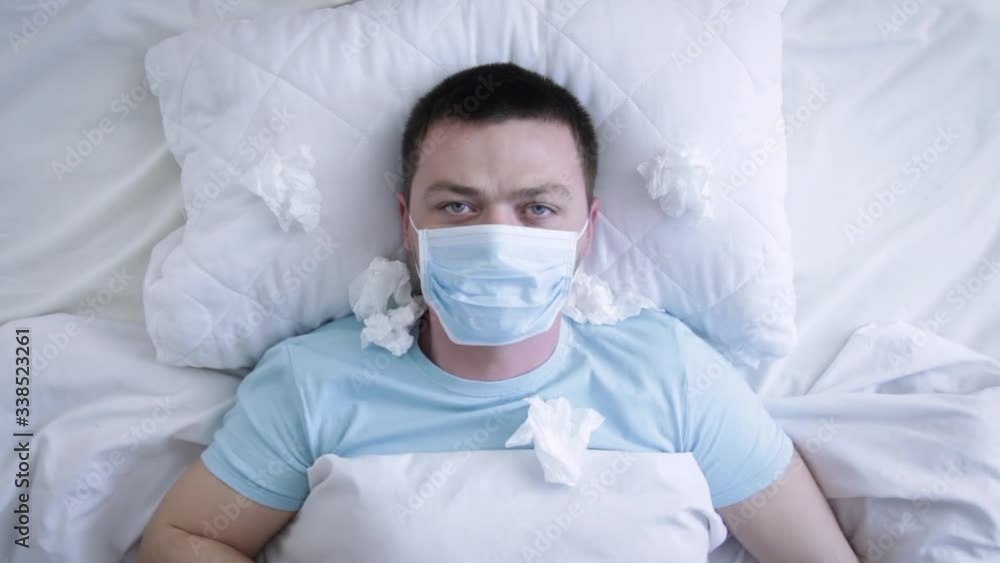 Man in medical mask coughing because of flu in the bed. Sick man with medical mask suffering from sore throat. Coronavirus Covid-19. Health concept Man in medical mask coughing because of flu in the bed. Sick man with medical mask suffering from sore throat. Coronavirus Covid-19. Health concept