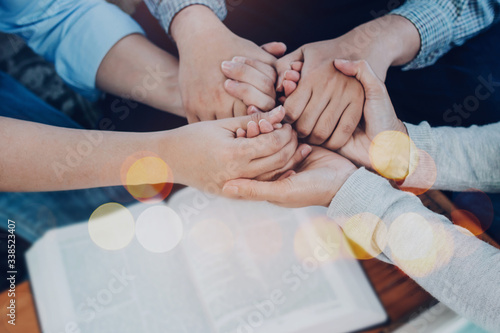 Close up of  people group holding hand and pray together over a blurred holy bible on wooden table, christian fellowship  or praying meeting in home concept with copy space photo