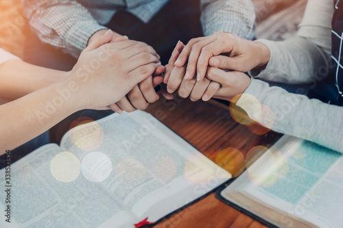 Close up of people group holding hand and pray together over a blurred holy bible on wooden table, christian fellowship or praying meeting in home concept with copy space