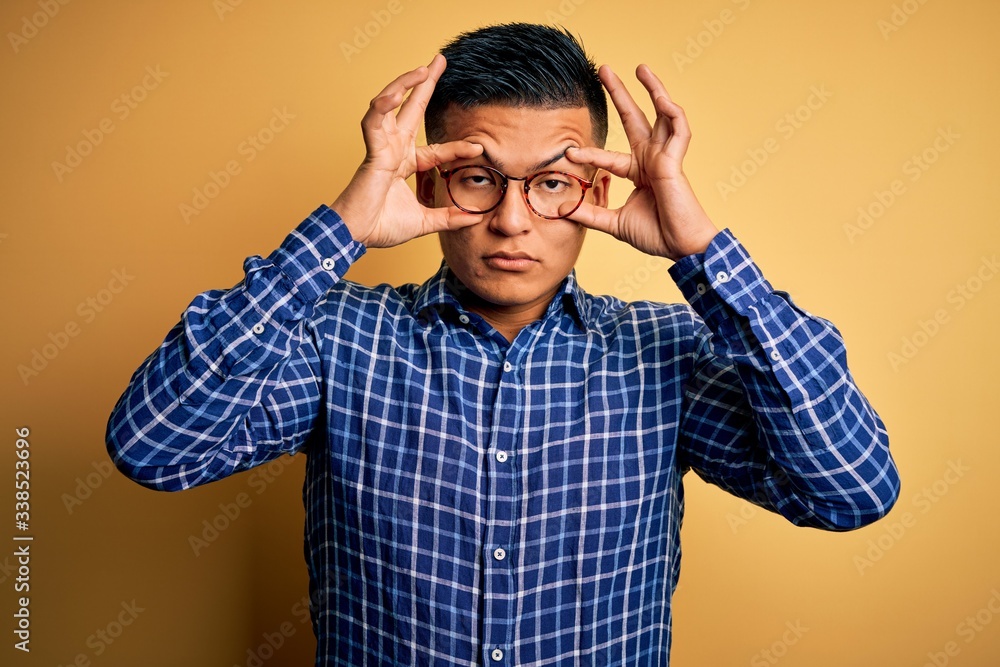 Young handsome latin man wearing casual shirt and glasses over yellow background Trying to open eyes with fingers, sleepy and tired for morning fatigue Young handsome latin man wearing casual shirt and glasses over yellow background Trying to open eyes with fingers, sleepy and tired for morning fatigue
