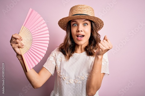 Young blonde fashion girl using wooden spanish hand fan on a hot summer day screaming proud and celebrating victory and success very excited, cheering emotion