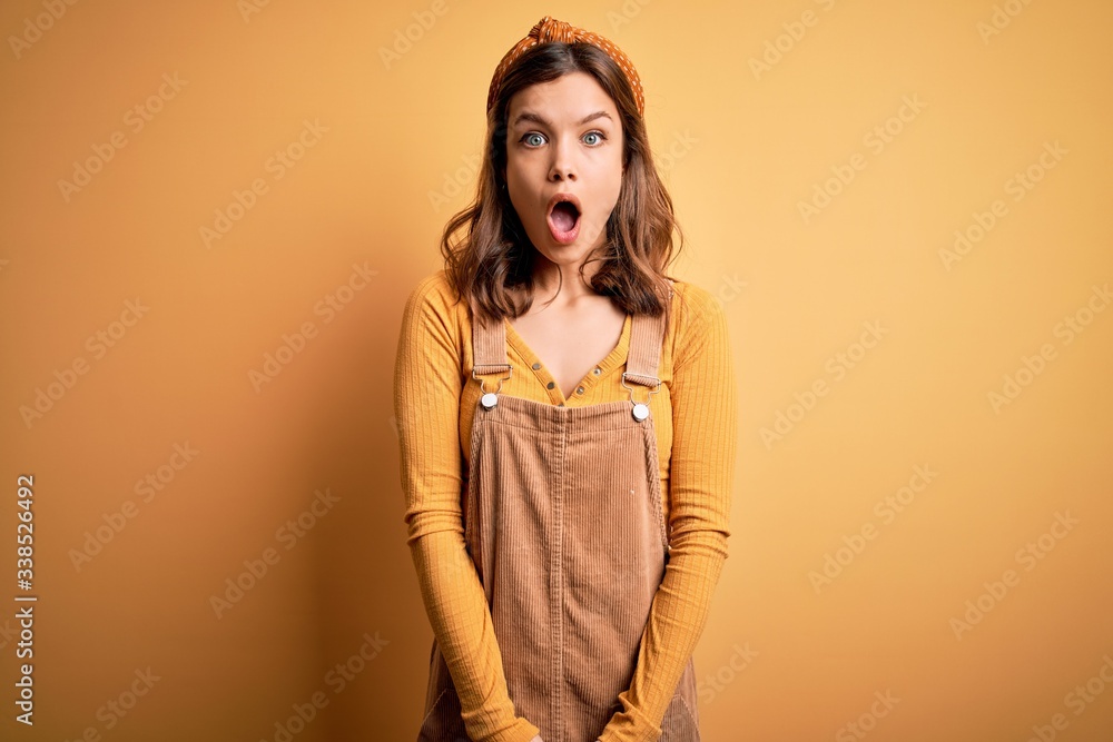 Young beautiful blonde girl wearing overall standing over yellow isolated background afraid and shocked with surprise and amazed expression, fear and excited face. Young beautiful blonde girl wearing overall standing over yellow isolated background afraid and shocked with surprise and amazed expression, fear and excited face.