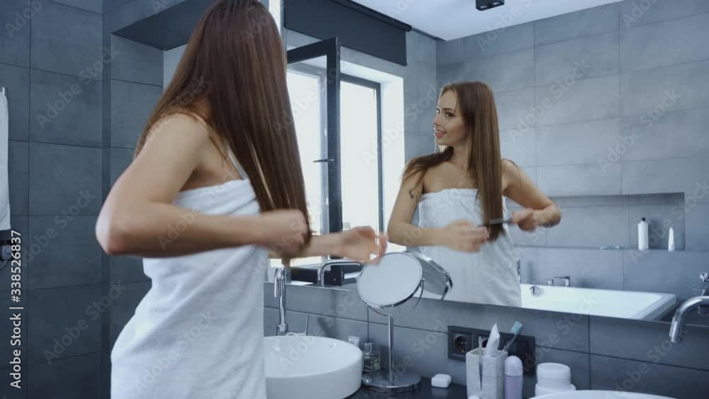 happy young woman combing hair in front of mirror in bathroom happy young woman combing hair in front of mirror in bathroom