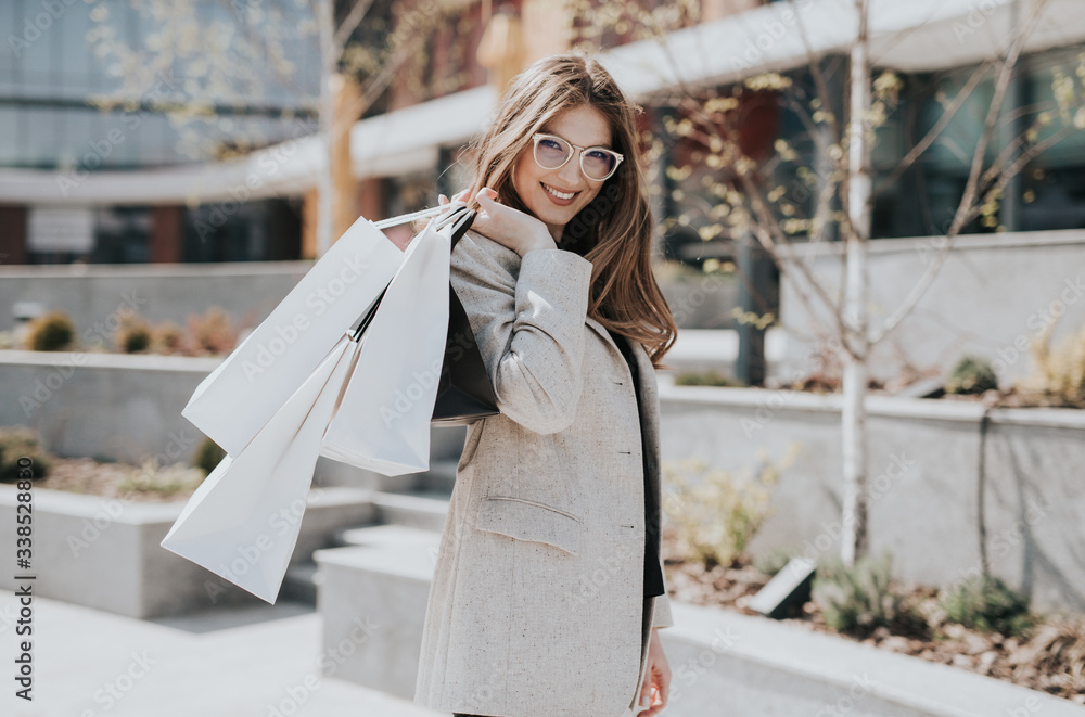 Fototapeta premium Attractive cute girl with the shopping bags and warm drink in her hand. Modern city megapolis on background. Lady order and shop online. Beautiful caucasian lady