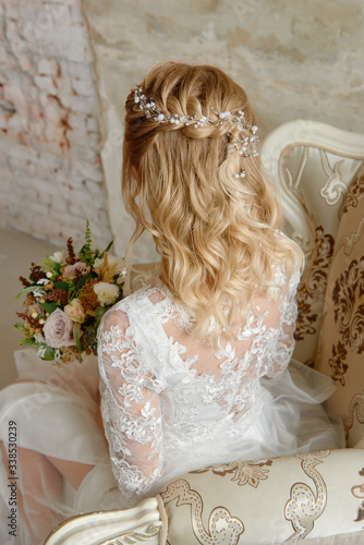 Beautiful bride wedding hairstyle with jewelry, back view. Blond girl with curly hair styling. Bride wirh wedding bouquet sitting on armchair