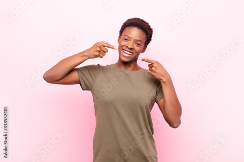 young pretty black womansmiling confidently pointing to own broad smile, positive, relaxed, satisfied attitude against pink wall photo