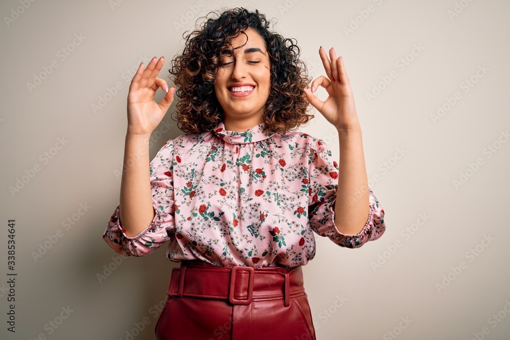 Young beautiful curly arab woman wearing floral t-shirt standing over isolated white background relax and smiling with eyes closed doing meditation gesture with fingers. Yoga concept. Young beautiful curly arab woman wearing floral t-shirt standing over isolated white background relax and smiling with eyes closed doing meditation gesture with fingers. Yoga concept.