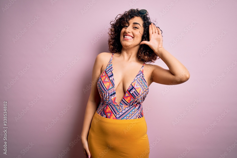 Young beautiful arab woman on vacation wearing swimsuit and sunglasses over pink background Waiving saying hello happy and smiling, friendly welcome gesture Young beautiful arab woman on vacation wearing swimsuit and sunglasses over pink background Waiving saying hello happy and smiling, friendly welcome gesture