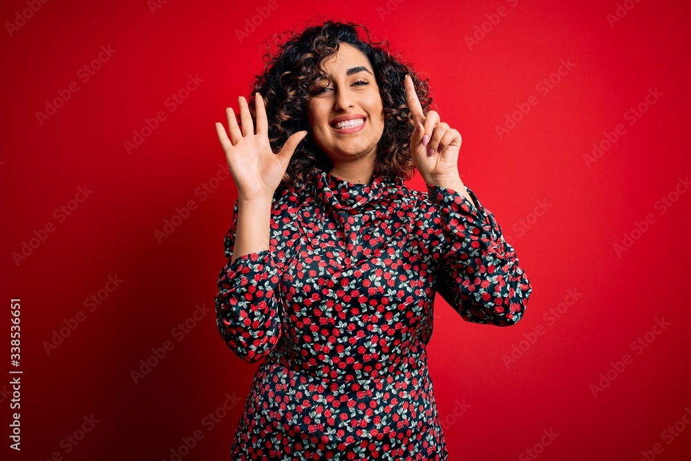 Young beautiful curly arab woman wearing casual floral dress standing over red background showing and pointing up with fingers number six while smiling confident and happy. Young beautiful curly arab woman wearing casual floral dress standing over red background showing and pointing up with fingers number six while smiling confident and happy.