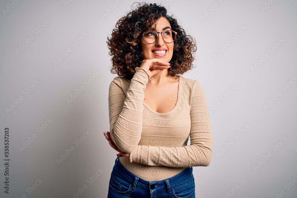 Young beautiful curly arab woman wearing casual t-shirt and glasses over white background with hand on chin thinking about question, pensive expression. Smiling and thoughtful face. Doubt concept.
