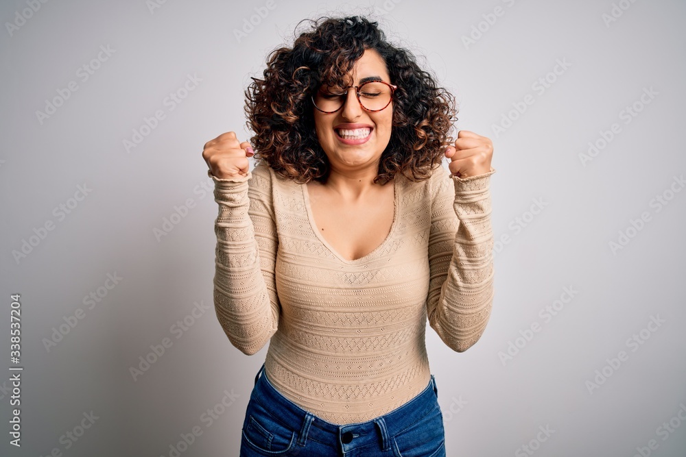 Young beautiful curly arab woman wearing casual t-shirt and glasses over white background excited for success with arms raised and eyes closed celebrating victory smiling. Winner concept. Young beautiful curly arab woman wearing casual t-shirt and glasses over white background excited for success with arms raised and eyes closed celebrating victory smiling. Winner concept.