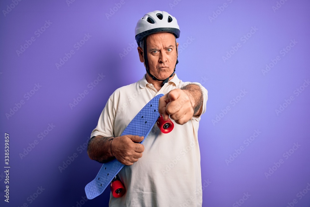 Middle age hoary man wearing security helmet holding skateboard over purple background pointing with finger to the camera and to you, hand sign, positive and confident gesture from the front Middle age hoary man wearing security helmet holding skateboard over purple background pointing with finger to the camera and to you, hand sign, positive and confident gesture from the front