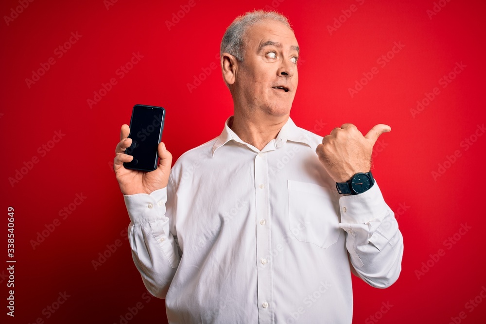 Middle age hoary man holding smartphone showing screen over isolated red background pointing and showing with thumb up to the side with happy face smiling Middle age hoary man holding smartphone showing screen over isolated red background pointing and showing with thumb up to the side with happy face smiling