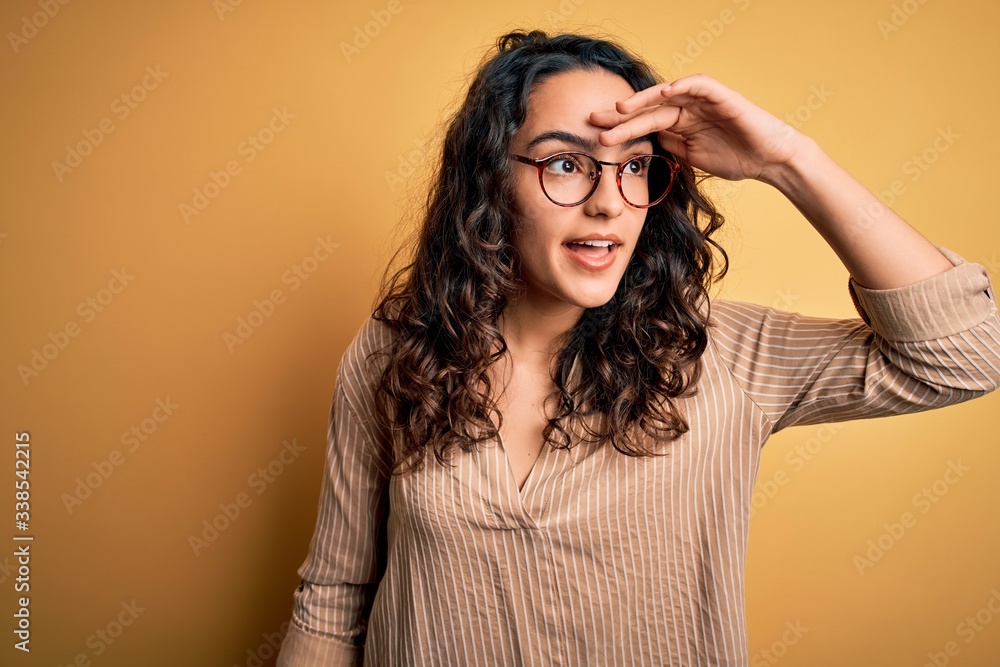 Beautiful woman with curly hair wearing striped shirt and glasses over yellow background very happy and smiling looking far away with hand over head. Searching concept. Beautiful woman with curly hair wearing striped shirt and glasses over yellow background very happy and smiling looking far away with hand over head. Searching concept.
