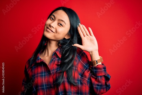 Young beautiful chinese woman wearing casual shirt over isolated red background Waiving saying hello happy and smiling, friendly welcome gesture © Krakenimages.com