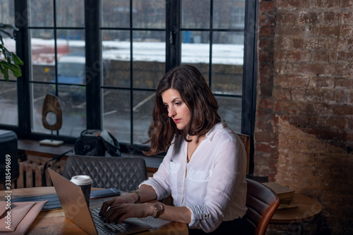 Business woman in white shirt working at laptop at home office near big window, drinking coffee. Chat on mobile. Wooden table, organizer.Remote work place. Quarantine of coronovirus pandemic covid-19