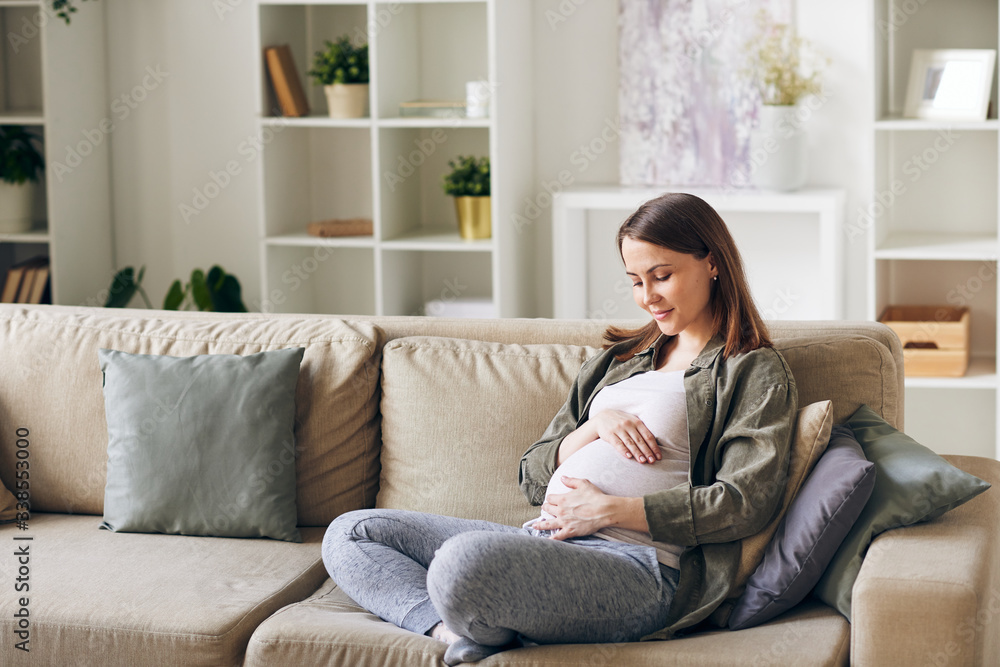 Calm attractive young pregnant woman sitting with crossed legs on sofa and embracing her belly Calm attractive young pregnant woman sitting with crossed legs on sofa and embracing her belly