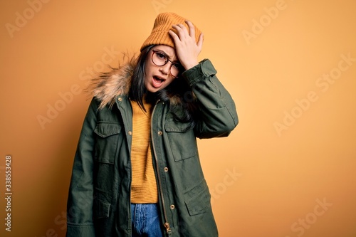Young brunette woman wearing glasses and winter coat with hat over yellow isolated background surprised with hand on head for mistake, remember error. Forgot, bad memory concept. © Krakenimages.com