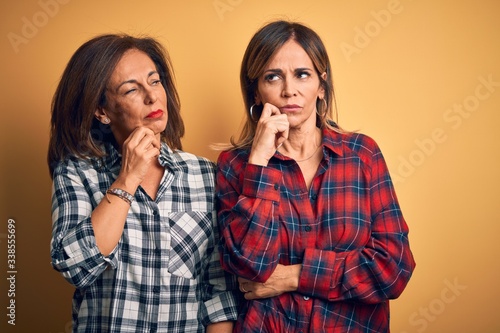 Middle age beautiful couple of sisters wearing casual shirt over isolated yellow background with hand on chin thinking about question, pensive expression. Smiling with thoughtful face. Doubt concept.
