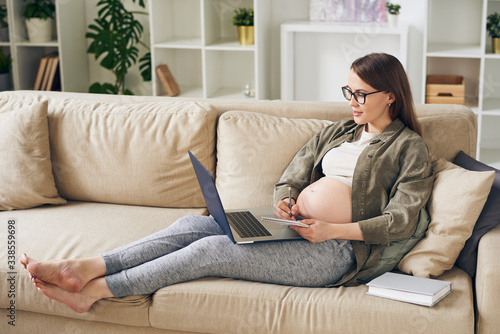 Attractive young pregnant woman in homewear sitting on sofa and reading article on laptop while making notes about maternity