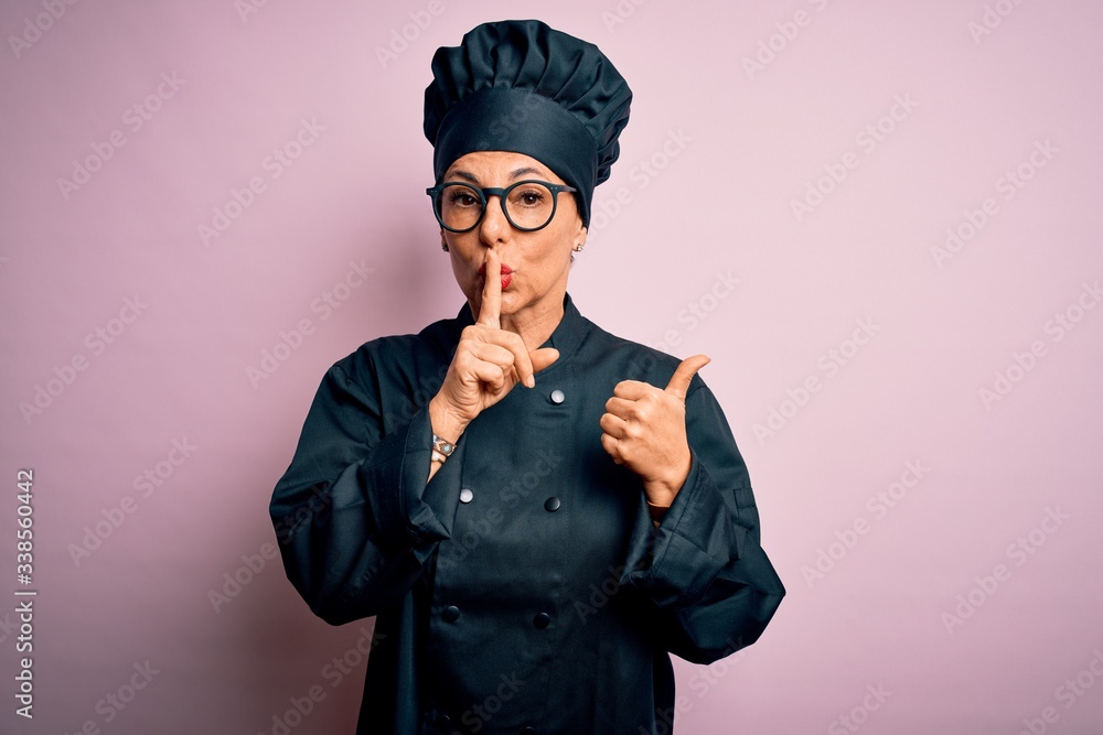Middle age brunette chef woman wearing cooker uniform and hat over isolated pink background asking to be quiet with finger on lips pointing with hand to the side. Silence and secret concept. Middle age brunette chef woman wearing cooker uniform and hat over isolated pink background asking to be quiet with finger on lips pointing with hand to the side. Silence and secret concept.