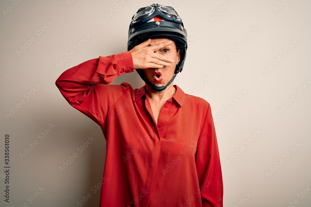 Middle age motorcyclist woman wearing motorcycle helmet over isolated white background peeking in shock covering face and eyes with hand, looking through fingers with embarrassed expression. Middle age motorcyclist woman wearing motorcycle helmet over isolated white background peeking in shock covering face and eyes with hand, looking through fingers with embarrassed expression.