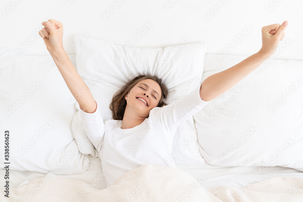 Young woman waking up late in morning on bed with white sheets and pillows, dressed in T-shirt, moving arms trying to get up, eyes closed, smiling Young woman waking up late in morning on bed with white sheets and pillows, dressed in T-shirt, moving arms trying to get up, eyes closed, smiling