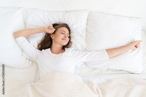 Young woman spending late morning in white bed under soft blanket, spreading and stretching arms to wake up, smiling with closed eyes