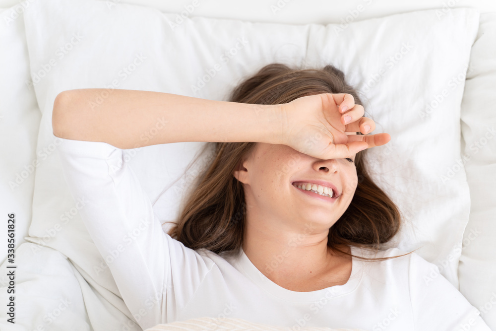 Fototapeta premium Close-up of young female showing sleepy smile while lying on bed on big white pillow, covering her eye and forehead with arm, not ready to get up