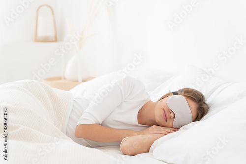 Young woman spending late morning in white bed, sleeping peacefully with eyes covered by gray eyemask, relaxing after long night
