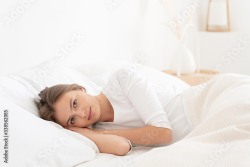 Young woman lying on white bed linen on one side, recently woke up, looking at camera with neutral face expression