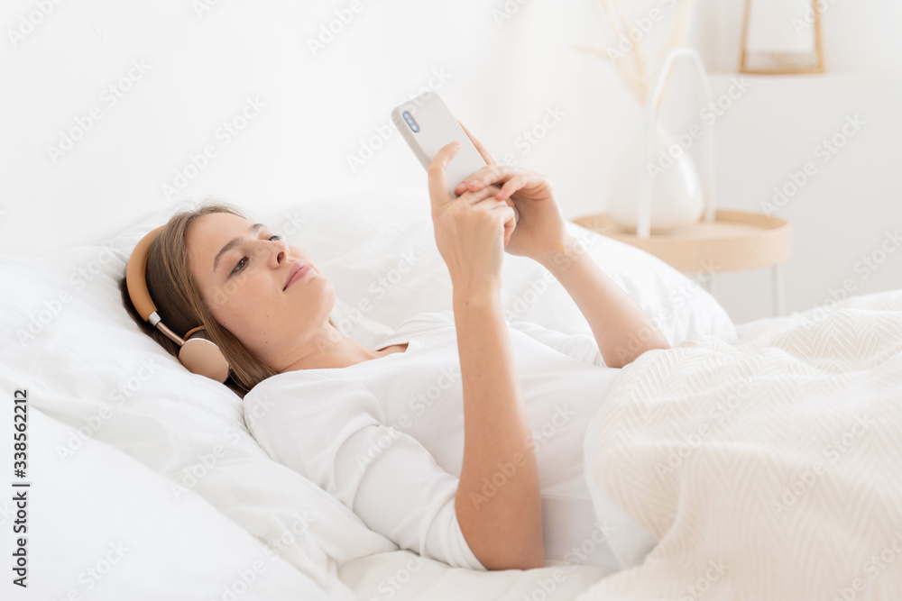 Young female watching video on smartphone from bed in morning, using wireless headphones to listen not to awake flatmates, enjoying free time Young female watching video on smartphone from bed in morning, using wireless headphones to listen not to awake flatmates, enjoying free time