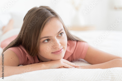 Close-up of young dreamy brunette girl, lying on white bed, looking aside as if thinking of future plans or date with her boyfriend
