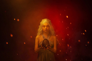 Art photo Portrait young beautiful woman blond hair in fire ash stained face. Fantasy power queen holding in hands egg mystic dragon. Backdrop mixed neon light red fiery sparks smoke. Halloween Image 