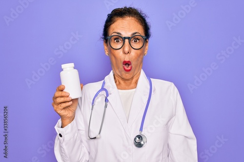 Middle age senior professional doctor woman holding pharmaceutical pills scared in shock with a surprise face, afraid and excited with fear expression