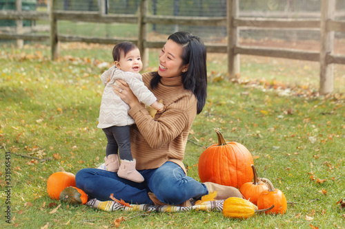 Happy smiling Asian Chinese mother with cute adorable baby. Mom and a daughter girl family sitting in autumn fall park outdoor with pumpkins. Halloween or Thanksgiving seasonal concept. © anoushkatoronto