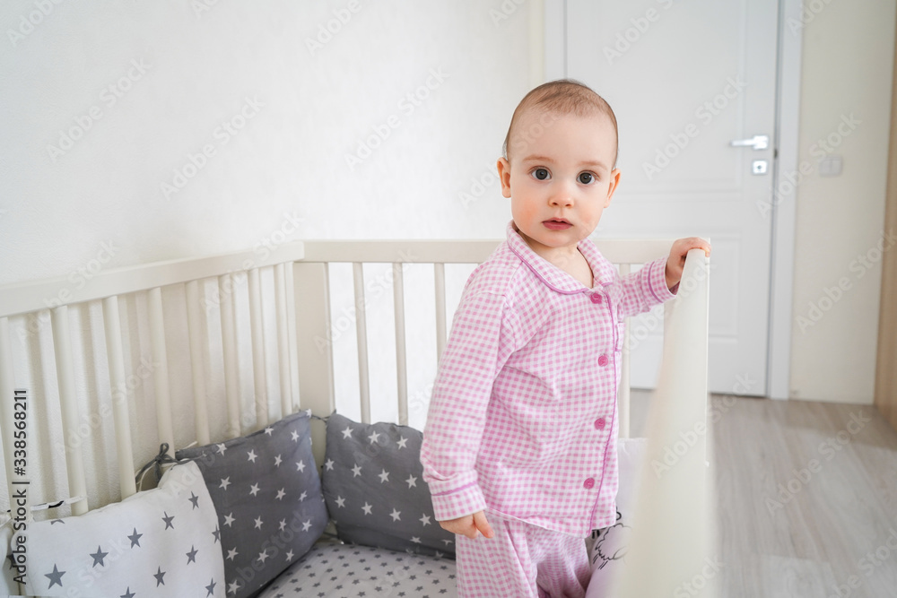 A small child in pink pajamas stands in the crib. The kid woke up and waits for his parents to get him out of the crib. A small child in pink pajamas stands in the crib. The kid woke up and waits for his parents to get him out of the crib.