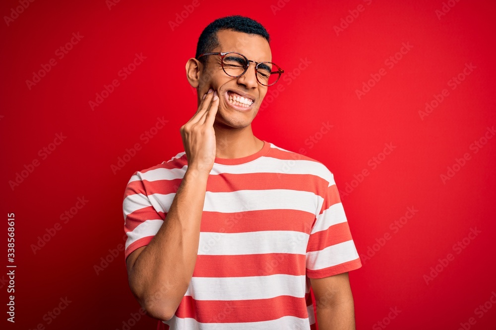Young handsome african american man wearing casual striped t-shirt and glasses touching mouth with hand with painful expression because of toothache or dental illness on teeth. Dentist Young handsome african american man wearing casual striped t-shirt and glasses touching mouth with hand with painful expression because of toothache or dental illness on teeth. Dentist